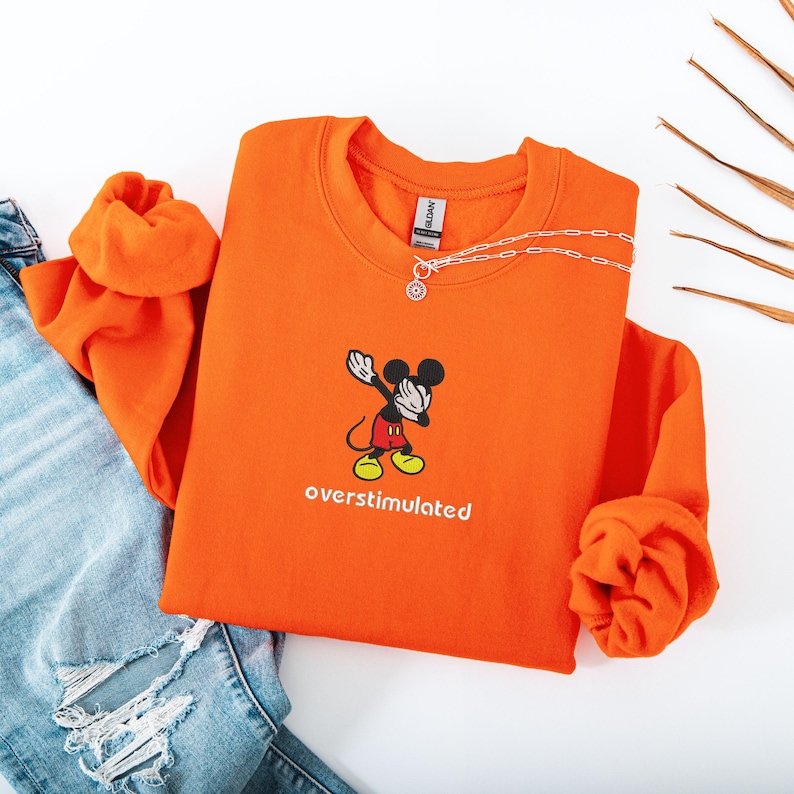 May include: An orange sweatshirt featuring a cartoon Mickey Mouse character dabbing, with the word "overstimulated" in white. A silver necklace is draped across the sweatshirt. The sweatshirt is folded next to a pair of blue jeans.
