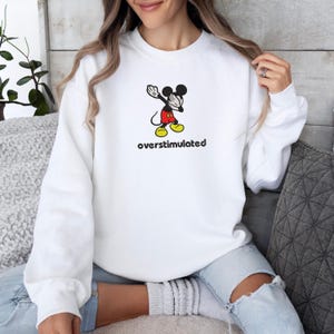 May include: White sweatshirt featuring a cartoon Mickey Mouse character in a dabbing pose. The character is in black, red, yellow, and white. The text "overstimulated" is printed below the character in black.
