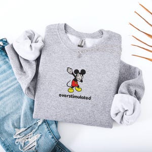 May include: A heather gray sweatshirt featuring a cartoon Mickey Mouse character in a dabbing pose, with the word "overstimulated" below. The sweatshirt is folded, with the sleeves visible. A silver necklace is draped across the top. Blue jeans are visible in the background.