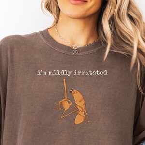 Op de afbeelding: Een bruine crewneck sweatshirt met de tekst "i'm mildly irritated" boven een cartoon garnalen illustratie. De garnaal steekt een middelvinger op. Het model draagt een zilveren ketting.