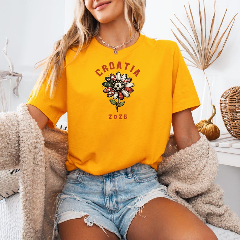 May include: A bright yellow short-sleeve t-shirt featuring a graphic of a flower with petals patterned in red and white checkers and green with white stars. The text CROATIA is printed above the flower and the number 2026 is printed below it.