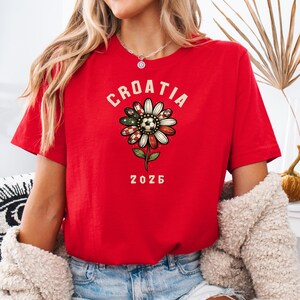 May include: A bright red short-sleeved t-shirt featuring a graphic of a daisy flower with petals patterned in red and white checkers and a soccer ball center. The text CROATIA is arched above the flower, and the number 2026 is printed below it.