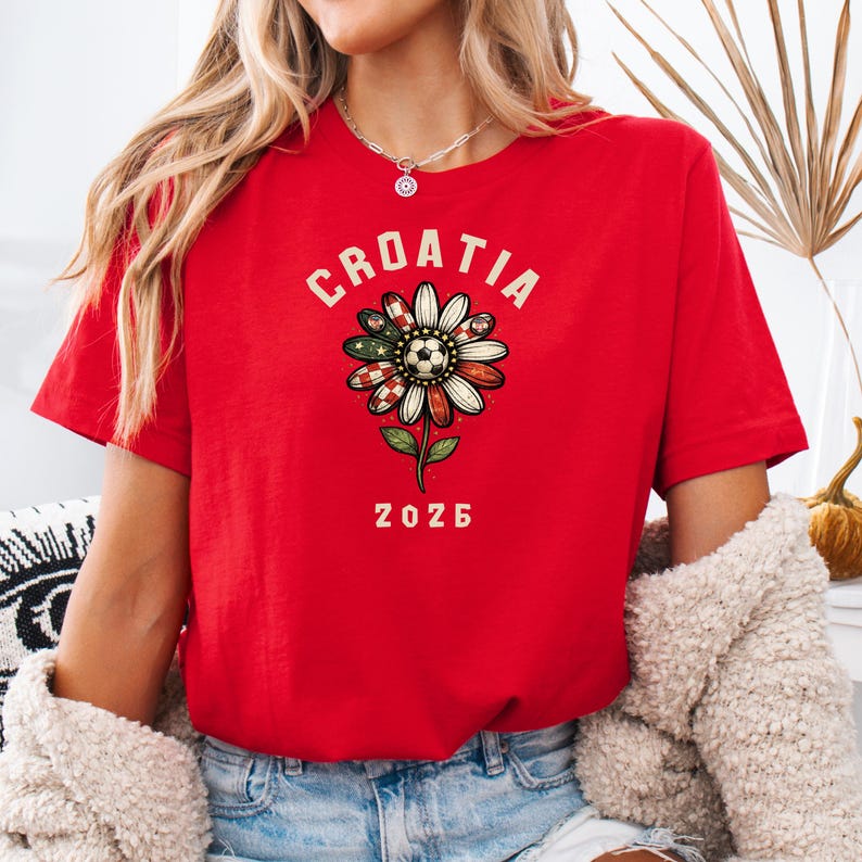 May include: A bright red short-sleeved t-shirt featuring a graphic of a daisy flower with petals patterned in red and white checkers and a soccer ball center. The text CROATIA is arched above the flower, and the number 2026 is printed below it.