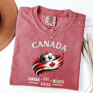 May include: A dusty rose Comfort Colors t-shirt featuring the word "CANADA" in a sparkly font above a soccer ball and a Canadian flag design. Below the design are the words "CANADA USA MEXICO 2026". A silver necklace with a pendant is draped on the shirt.