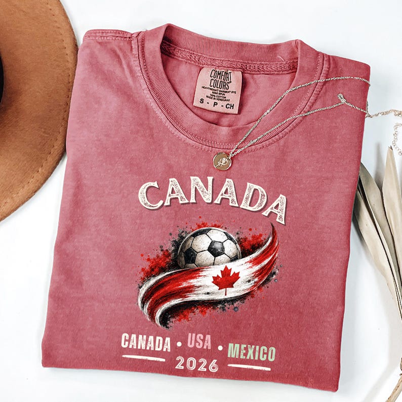 May include: A dusty rose Comfort Colors t-shirt featuring the word "CANADA" in a sparkly font above a soccer ball and a Canadian flag design. Below the design are the words "CANADA USA MEXICO 2026". A silver necklace with a pendant is draped on the shirt.
