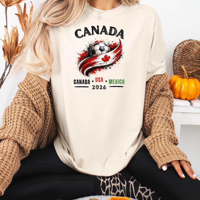 May include: Cream-colored t-shirt featuring a soccer ball design with the word "CANADA" above it. The design includes a red and white swoosh with a maple leaf and the text "CANADA USA MEXICO 2026".