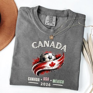 May include: Gray t-shirt featuring the word "CANADA" above a soccer ball and a red and white Canadian flag design. Below the design are the words "CANADA USA MEXICO 2026". A silver necklace is draped across the shirt.