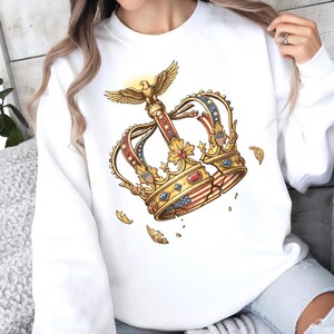 May include: A white long-sleeve sweatshirt featuring a central graphic of a gold crown adorned with red, white, and blue accents, including small American flag patterns and a golden eagle perched on top, with scattered gold decorative fragments.