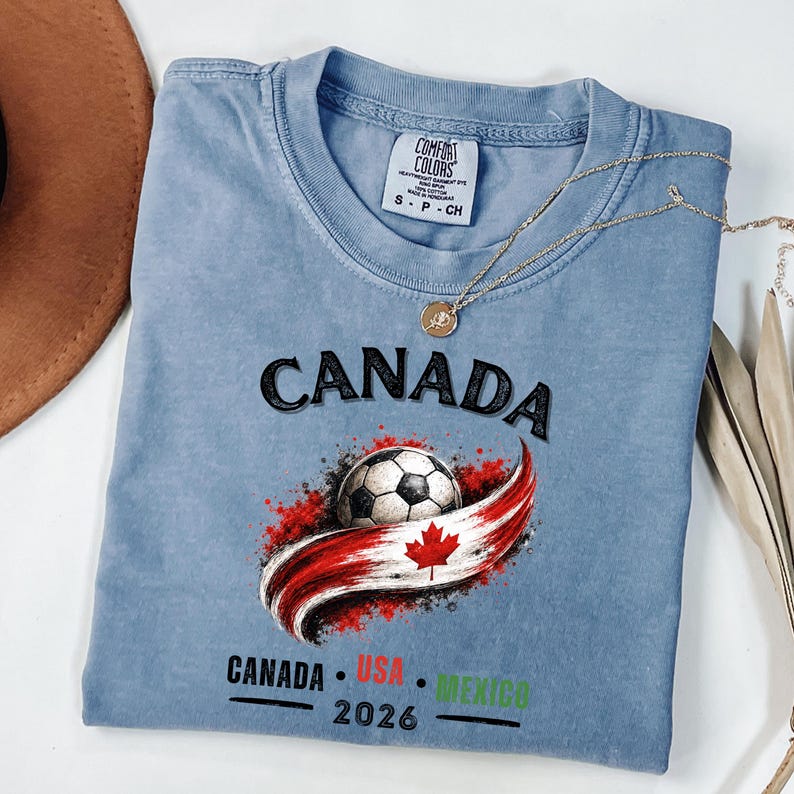 May include: A light blue t-shirt with the word "CANADA" in black above a soccer ball and a red and white design. Below the design are the words "CANADA USA MEXICO 2026". The shirt has a Comfort Colors tag.