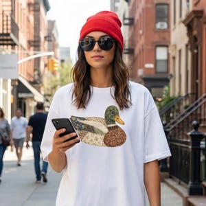 May include: A white short-sleeved t-shirt featuring a graphic of a duck composed of various brown, green, and cream-colored floral and patterned fabric patches. The shirt is worn with a bright red knit beanie and dark-tinted sunglasses.