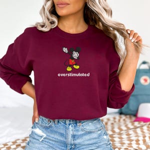 May include: A maroon sweatshirt featuring a cartoon Mickey Mouse character in a dabbing pose. The text "overstimulated" is printed in white below the graphic. The sweatshirt is a solid color and has a crew neck.