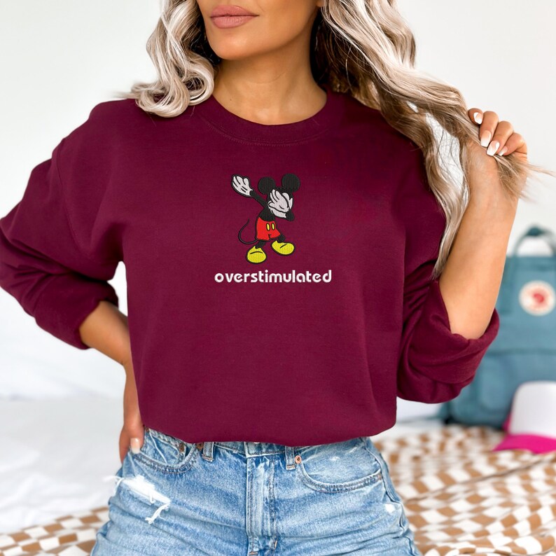 May include: A maroon sweatshirt featuring a cartoon Mickey Mouse character in a dabbing pose. The text "overstimulated" is printed in white below the graphic. The sweatshirt is a solid color and has a crew neck.