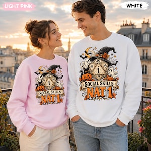 May include: A light pink sweatshirt and a white sweatshirt, each featuring a graphic print of a twenty-sided die wearing a witch hat, surrounded by ghosts, pumpkins, bats, and spiderwebs, with the text "SOCIAL SKILLS: NAT 1" printed below.