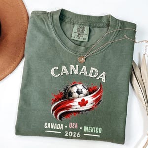 May include: An olive green t-shirt with the word "CANADA" in white block letters. Below is a soccer ball with a red and white Canadian flag design. The text "CANADA USA MEXICO 2026" is printed below the flag.