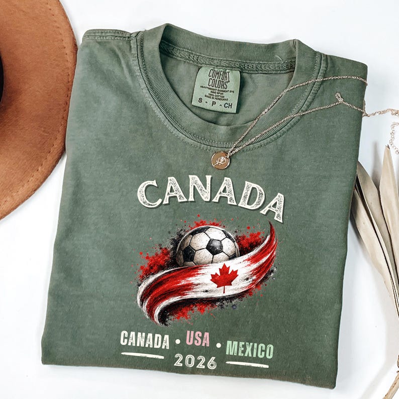 May include: An olive green t-shirt with the word "CANADA" in white block letters. Below is a soccer ball with a red and white Canadian flag design. The text "CANADA USA MEXICO 2026" is printed below the flag.