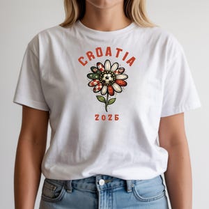 May include: A white t-shirt featuring a graphic of a flower with petals patterned in red and white checkers and green accents. A soccer ball sits at the center of the flower. The text CROATIA is arched above the flower, and 2026 is printed below.