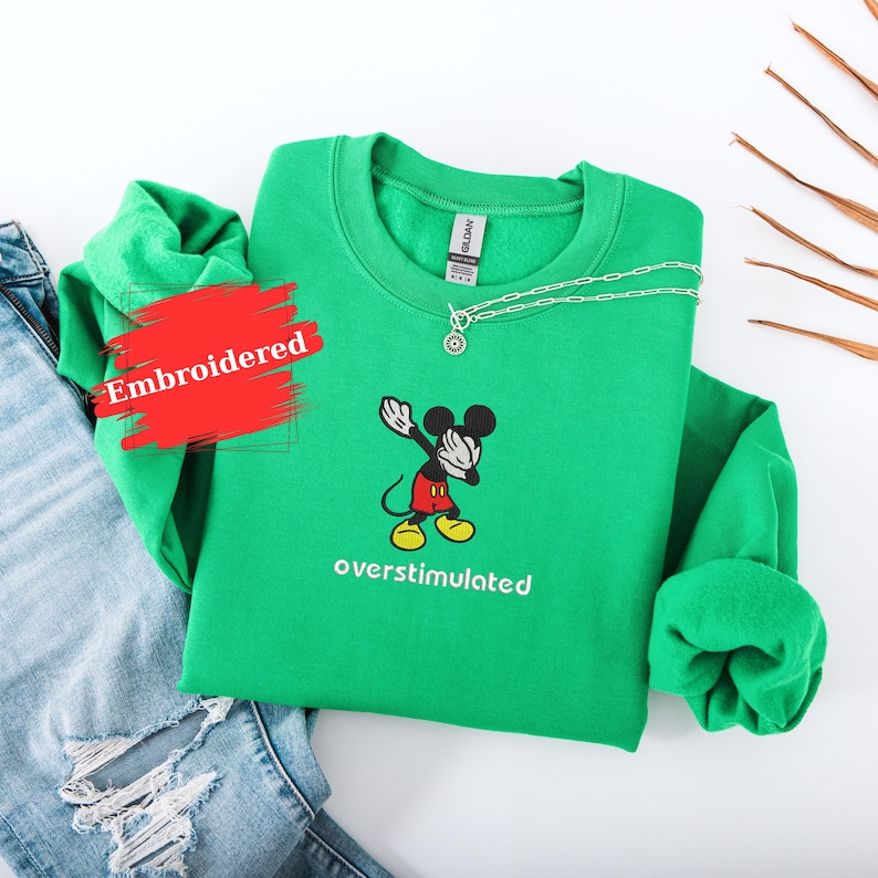 May include: A green embroidered sweatshirt featuring a cartoon Mickey Mouse character in a dabbing pose. The text "overstimulated" is below the character. A silver necklace is draped on the sweatshirt. Blue jeans are visible in the background.