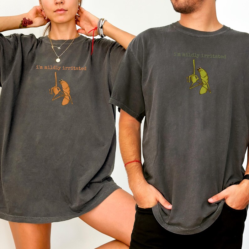 Op de afbeelding: Twee donkergrijze t-shirts met de tekst "I'm mildly irritated" boven een cartoonafbeelding van een persoon die een plant water geeft. De afbeelding is in bruin- en groentinten. De shirts zijn korte mouwen.