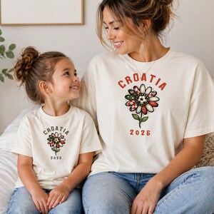 May include: Two cream-colored short-sleeve t-shirts featuring a graphic of a daisy flower with a soccer ball center. The petals are patterned with red and white checkers and green accents. The text CROATIA and 2026 is printed in red.