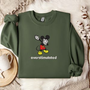 May include: An olive green sweatshirt featuring a cartoon Mickey Mouse character in a dabbing pose. The character is wearing red shorts and yellow shoes. The word "overstimulated" is printed in white below the character.