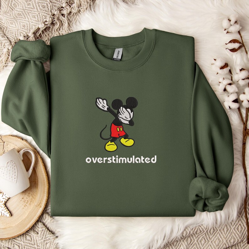 May include: An olive green sweatshirt featuring a cartoon Mickey Mouse character in a dabbing pose. The character is wearing red shorts and yellow shoes. The word "overstimulated" is printed in white below the character.