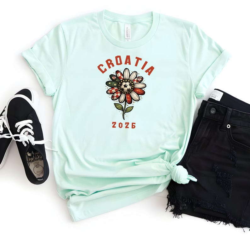 May include: A light mint green t-shirt featuring a graphic of a flower with a soccer ball center and red and white checkered petals. The text CROATIA is arched above the flower, and the number 2026 is printed below in a bold red font.