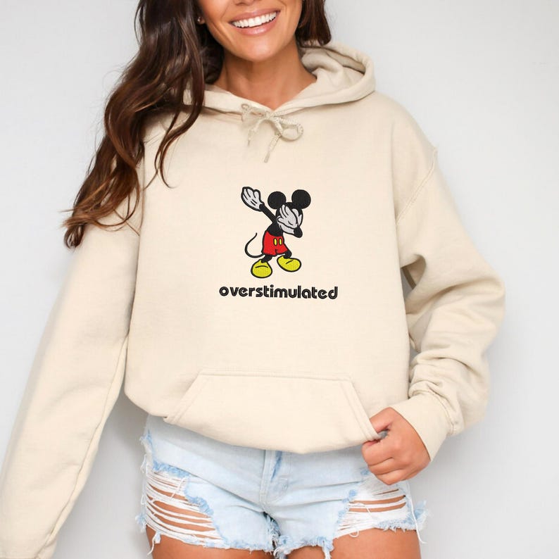 May include: Beige hoodie with a front pocket and drawstring. Features a graphic of Mickey Mouse dabbing with the word "overstimulated" below. The hoodie is a neutral color and has a relaxed fit.