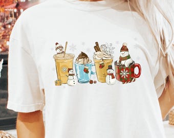 May include: A white short-sleeve t-shirt featuring a graphic print of four beverage containers, including cups and mugs, decorated with snowmen, snowflakes, small stockings, and holiday-themed elements like cinnamon sticks and a candy cane.