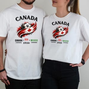 May include: White t-shirts with a soccer-themed graphic. The design features the word "CANADA" above a soccer ball and a red and white ribbon with a maple leaf. Below, it reads "CANADA &bull; USA &bull; MEXICO 2026".