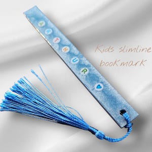 Puede incluir: Un marcapáginas azul claro y delgado con el nombre "ARTHUR" escrito en letras coloridas. El marcapáginas tiene una borla azul adjunta y el texto "Kids slimline bookmark".