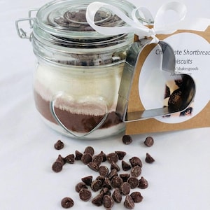 May include: A glass jar containing layered ingredients for chocolate shortbread biscuits, with a heart-shaped design. A metal heart-shaped cookie cutter and a tag with text are attached. Chocolate chips are scattered around the jar.