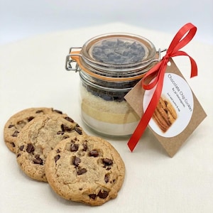 May include: A glass jar containing layered ingredients for chocolate chip cookies, tied with a red ribbon and tag. Three baked chocolate chip cookies are in front of the jar. The tag reads "Chocolate Chip Cookies".