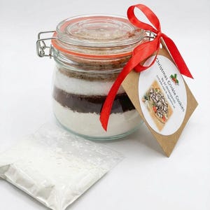 May include: A glass jar containing layered ingredients for Christmas Crinkle Cookies, secured with a red ribbon and a tag. A small plastic bag of white powder is in front of the jar. The jar has a metal clasp and a red rubber seal.