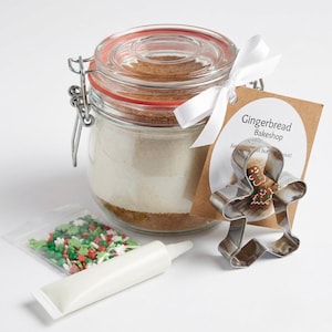 May include: A gingerbread baking kit featuring a glass jar with dry ingredients, a gingerbread man cookie cutter, sprinkles, and a tube of icing. The kit includes a tag with the text "Gingerbread Bakeshop". A white ribbon is tied around the jar.