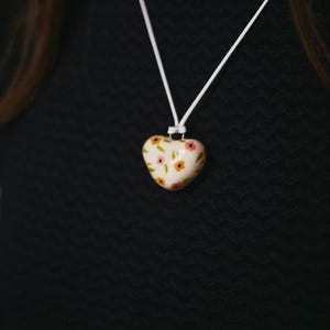 Op de afbeelding: Een hartvormige hangerketting met een wit koord. De hanger is crèmekleurig en versierd met kleine roze en gele bloemmotieven. De achtergrond is een zwarte, getextureerde ondergrond.