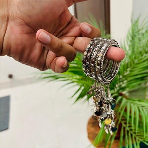 Può includere: Braccialetti color argento con decorazioni e ciondoli pendenti. I ciondoli presentano piccole forme di cuore e foglie in vari colori. I braccialetti sono presentati su uno sfondo di piante verdi.