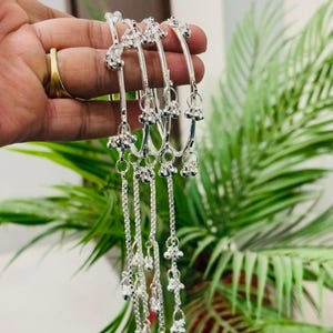 May include: Silver anklets with dangling charms and delicate chain details. The anklets are held in a hand, showcasing their intricate design and reflective surface. The charms and chains add movement and visual interest to the jewellery.