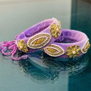 Puede incluir: Dos brazaletes de terciopelo lavanda con adornos de cuentas doradas y blancas. Cada brazalete presenta detalles de flores doradas y diseños ovalados con cuentas. Una hebra de cuentas moradas cuelga de un brazalete.