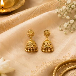 Könnte beinhalten: Ein Paar goldfarbene Jhumka-Ohrringe. Jeder Ohrring hat ein florales Design an der Oberseite, einen glockenförmigen Körper mit komplizierten Mustern und kleine, baumelnde Perlen an der Unterseite. Die Ohrringe liegen auf einem cremefarbenen Stoff.