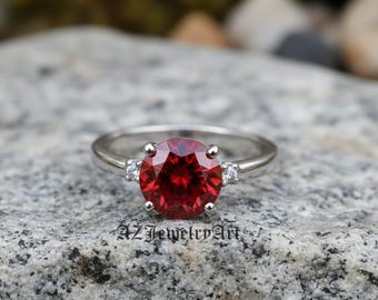 May include: A silver ring with a large, round, faceted red gemstone as the centerpiece. Two smaller white gemstones flank the main stone. The ring is set on a gray stone surface. The text "AZ Jewelry Art" is visible.