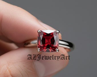 May include: A silver ring featuring a large, square-cut, deep red gemstone. The ring's band is smooth and polished, and the gemstone is held in place by four prongs. The text "AZJewelryArt" is visible on the ring.