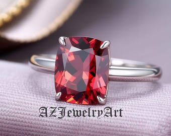 May include: A silver ring featuring a large, cushion-cut, deep red gemstone. The gemstone is set with four prongs and the band is smooth and polished. The text "AZJewelryArt" is printed on the surface.