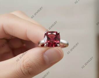 May include: A silver ring with a large, cushion-cut, deep red gemstone. The ring is held up against a neutral background, showcasing the gemstone's facets and the ring's simple band. The ring is a piece of jewelry.