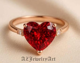 May include: Rose gold ring featuring a large, heart-shaped, faceted red gemstone. The ring has small accent stones on either side of the heart and the text "AZJewelryArt" at the bottom.
