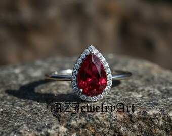 May include: A pear-shaped, ruby-red gemstone ring with a halo of small, clear diamonds. The ring is set on a silver band and rests on a gray stone surface. The text "AZ Jewelry Art" is visible.