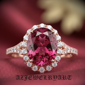 Könnte beinhalten: Roségoldener Ring mit einem großen, oval geschliffenen, tiefroten Edelstein, umgeben von kleinen, klaren Edelsteinen. Das Band ist ebenfalls mit kleineren Edelsteinen besetzt. Der Ring befindet sich vor einem bordeauxroten Hintergrund.