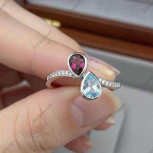 May include: A sterling silver ring featuring two pear-shaped gemstones. One is a deep red, the other a light blue. The band is adorned with small, clear accent stones. The ring is a piece of jewelry.