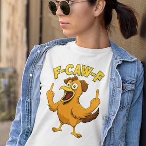 Könnte beinhalten: Weißes T-Shirt mit einem Cartoon-Hühner-Motiv. Das Huhn ist braun und gelb und zeigt mit beiden Händen den Mittelfinger. Der Text "F-CAW-F" steht in gelben Buchstaben über dem Huhn. Die Person trägt eine Jeansjacke.