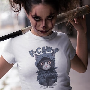Könnte beinhalten: Weißes T-Shirt mit einem Cartoon-Kätzchen in einem dunkelblauen Kapuzenmantel und dem Text "F-CAW-F". Die Person im Bild hält einen Baseballschläger. Die Person hat Clown-Make-up.