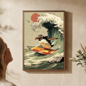 Könnte beinhalten: Ein gerahmter Kunstdruck zeigt einen Papageientaucher, der auf einer großen Welle unter einer roten Sonne surft. Das Kunstwerk ist im Vintage-Stil mit einem braunen Rahmen gehalten. Der Druck wird an einer beigefarbenen Wand gezeigt, mit einer Frau, die das Kunstwerk betrachtet.